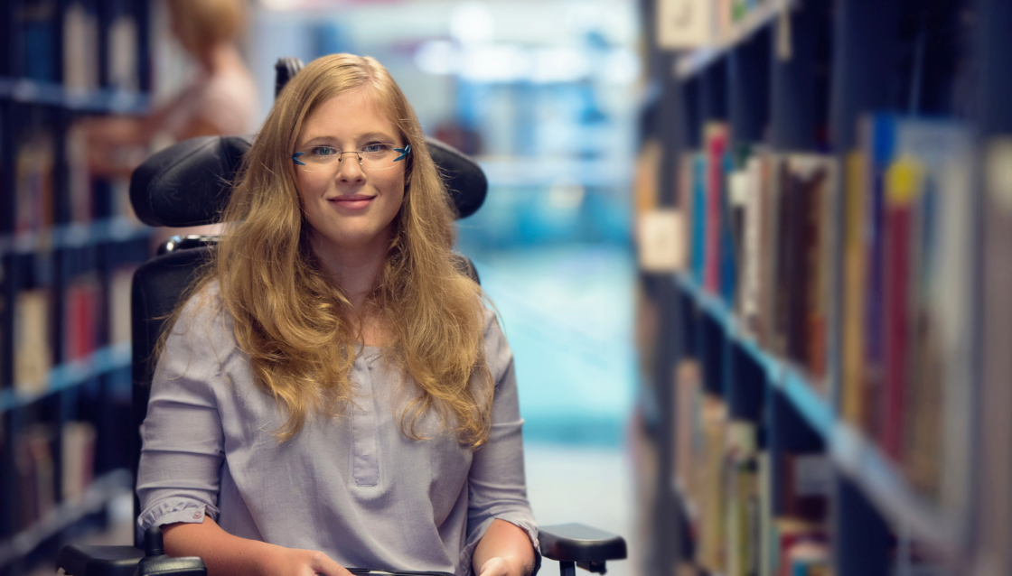 Image of young woman in electric wheelchair inside a library holding a tablet device and smiling at the camera.